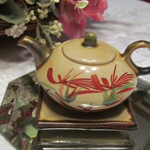 May include: A small, beige teapot with a lid and a brown handle. The teapot is decorated with red and green floral designs. The teapot is sitting on a brown and beige square coaster.