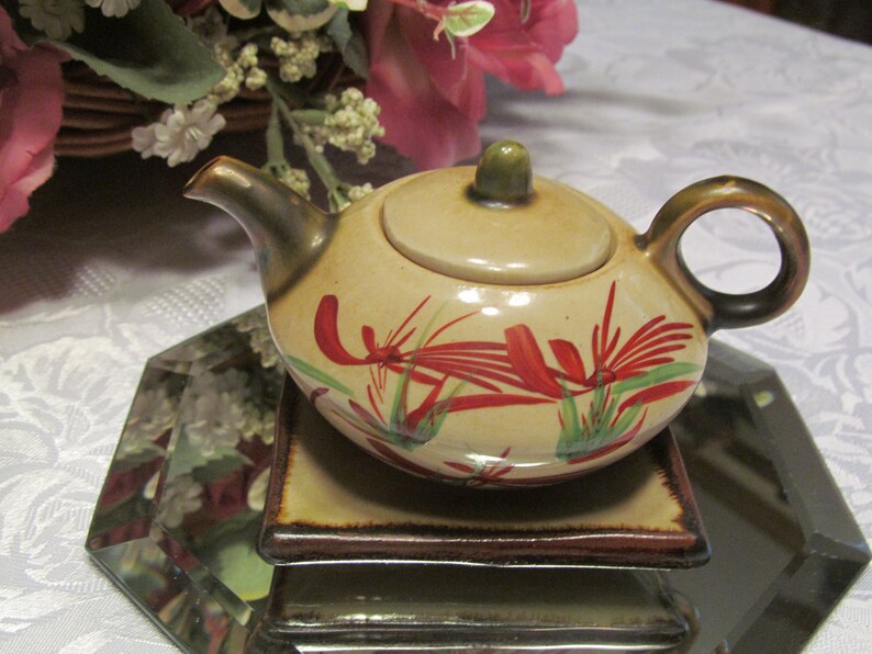 May include: A small, beige teapot with a lid and a brown handle. The teapot is decorated with red and green floral designs. The teapot is sitting on a brown and beige square coaster.