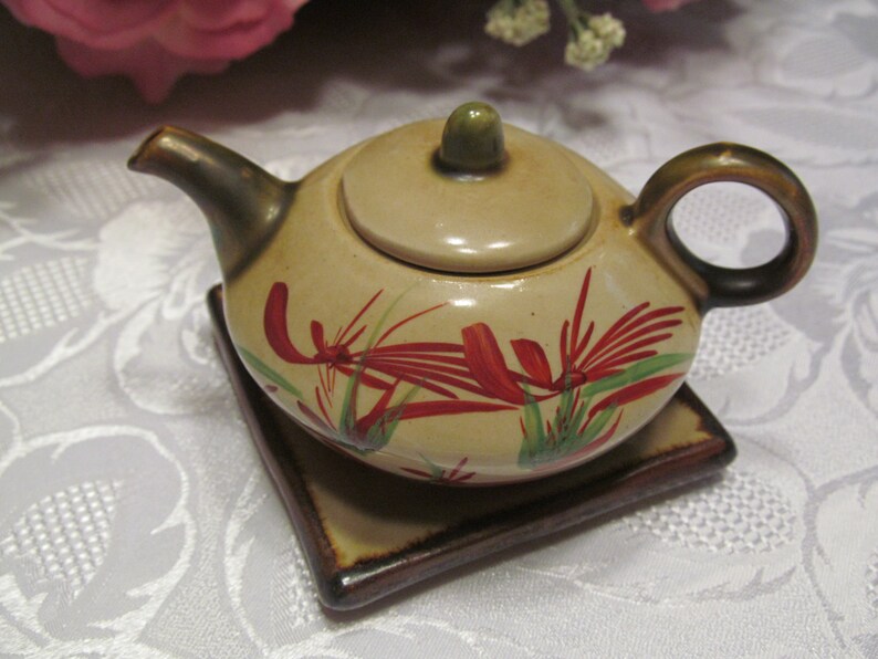 May include: A small, beige teapot with a lid and a brown handle. The teapot has a floral design with red and green accents. The teapot is sitting on a brown square coaster.