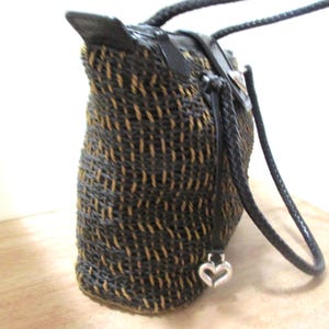 May include: A woven tote bag with a black and tan pattern. The bag has braided black handles and leather accents. A silver heart charm hangs from the side. The bag is made of a woven material.