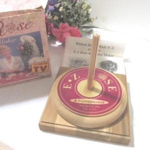 May include: A wooden E-Z Rose & Flower Maker with a red and gold label. The label reads "E.Z Rose & Flower Maker" and "Made in USA". The box in the background reads "Rose Maker" and "As Seen on TV".