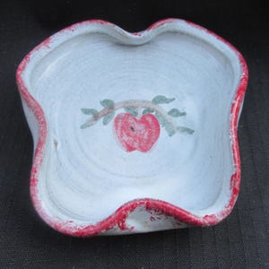 May include: A small, white ceramic bowl with a wavy rim, painted with a red apple on a branch. The rim is painted red, and the bowl has a rustic, handmade appearance. The bowl is likely used for serving small portions of food or as a decorative item.