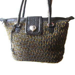 May include: Woven tote bag with black and gold threads, featuring a black leather flap with a silver heart-shaped emblem. The bag has braided black handles and a zippered top closure. A stylish accessory for everyday use.