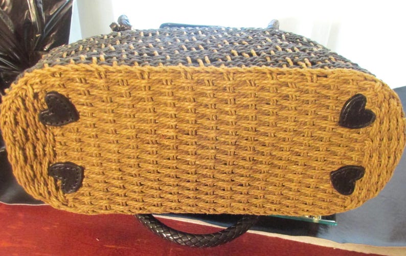 May include: Woven tote bag with a two-tone design. The top section is dark brown, and the bottom is tan. The base features four black heart-shaped accents. The bag has a black braided handle.