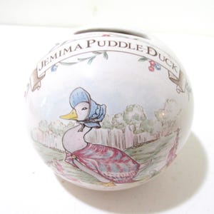 May include: A white ceramic Jemima Puddle-Duck money bank. The bank features a detailed illustration of Jemima Puddle-Duck in a blue bonnet and patterned dress, set against a pastoral background. The name "Jemima Puddle-Duck" is written in a banner around the top.