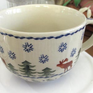 May include: A cream-colored ceramic mug featuring a hand-painted winter scene with green pine trees, brown reindeer, and blue snowflakes. The rim is decorated with a blue wavy line pattern, and the mug includes a single side handle.