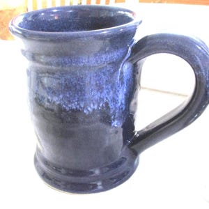 May include: A handmade ceramic mug with a dark blue glaze. The mug has a large handle and a slightly flared rim. The glaze transitions from a lighter blue at the top to a darker shade at the base, creating a textured appearance.