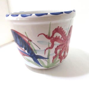 May include: A white ceramic planter with a blue scalloped rim. The planter is decorated with a red octopus and a blue and red fish design, with green seaweed accents. The design is hand-painted.