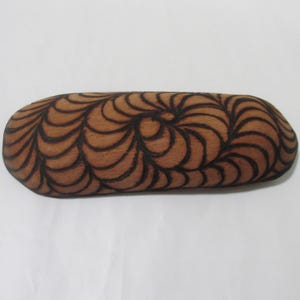 May include: A wooden eyeglasses case with a brown finish and a swirling, black, patterned design. The case is oval-shaped and appears to be made of a single piece of wood. The design is symmetrical and covers the entire surface.