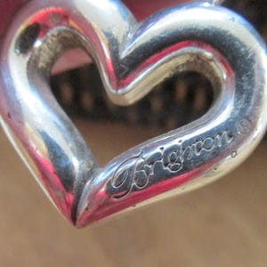 May include: A close-up of a silver heart-shaped pendant. The pendant has a hollow center and the word "Brighton" is engraved on the lower right side. The pendant is on a chain.
