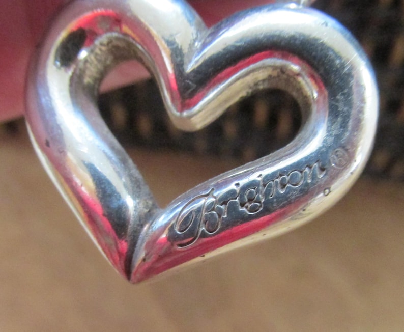 May include: A close-up of a silver heart-shaped pendant. The pendant has a hollow center and the word "Brighton" is engraved on the lower right side. The pendant is on a chain.