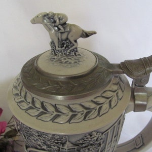 May include: A ceramic beer stein with a detailed design of a horse and rider. The lid features a sculpted horse and rider, and the stein has a handle with a decorative metal piece. The stein is decorated with a leafy pattern.