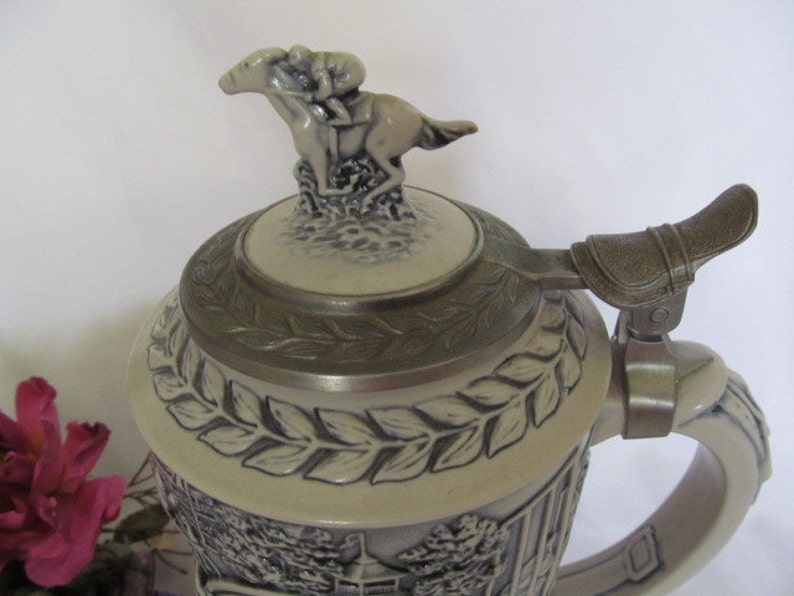 May include: A ceramic beer stein with a detailed design of a horse and rider. The lid features a sculpted horse and rider, and the stein has a handle with a decorative metal piece. The stein is decorated with a leafy pattern.
