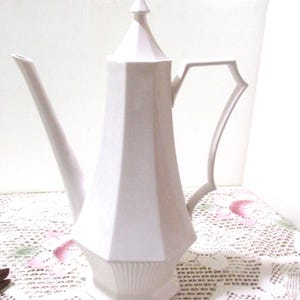 May include: A white, geometric-shaped ceramic coffee pot with a long spout and a handle. The pot has a pointed top and a decorative base. The design is a light, neutral color, suitable for serving coffee or tea.