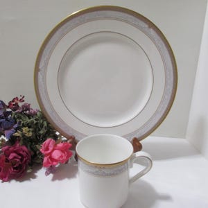 May include: White teacup and plate set with gold trim and a decorative border. The plate is large and round, and the teacup has a handle. The set is displayed on a white surface with flowers in the background. A classic tableware set.