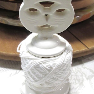 May include: A white ceramic yarn holder featuring a stylized cat head topper with cut-out eyes and mouth. The base is a circular pedestal designed to hold a spool of white yarn, which is wrapped around the central column beneath the cat head.