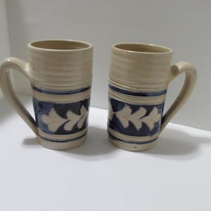 May include: Two tall cylindrical ceramic mugs with a light beige base color. Each mug features a wide horizontal band of deep blue glaze around the center, decorated with a repeating white leaf-like pattern. Both mugs have a curved handle.