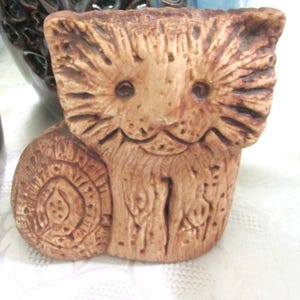 May include: A small, light brown, textured clay figurine of a cat. The cat has a wide, flat face with etched lines representing fur, circular eyes, and a simple smile. The body features carved circular and vertical patterns on its surface.