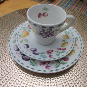 May include: A white ceramic cup and saucer set featuring a colorful fruit pattern. The design includes illustrations of purple grapes, red cherries, yellow pears, and green leaves. A single cherry motif is painted on the interior of the cup.