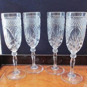 May include: Four clear crystal champagne flutes with intricate diamond and leaf patterns. Each flute has a round base, a slender stem with a decorative bead, and a tall, textured bowl. The glasses are arranged on a wooden surface.