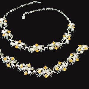May include: A silver tone necklace set with two strands of linked, stylized flower-shaped pendants. Each pendant features a cluster of orange rhinestones.