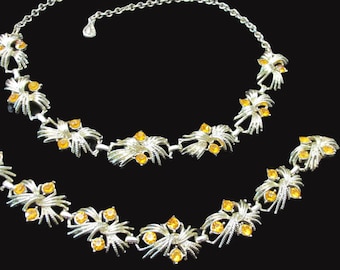 May include: A silver tone necklace set with two strands of linked, stylized flower-shaped pendants. Each pendant features a cluster of orange rhinestones.