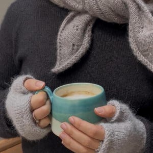 May include: A person wearing a gray knitted scarf, sweater, and fingerless gloves holds a teal and white mug of coffee. The mug has a handle and is filled with a creamy beverage. The gloves and scarf are a light gray color.