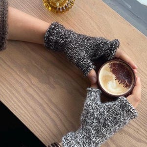 May include: A pair of gray and white knit fingerless gloves. The gloves are worn while holding a cup of coffee with a foamy top. A lit candle in a decorative holder sits on a wooden table.