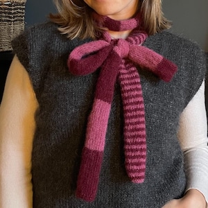 May include: A knitted scarf in shades of pink and burgundy, tied in a bow. The scarf has a striped pattern on the ends and is worn over a gray sleeveless sweater.