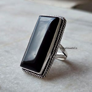 Könnte beinhalten: Ein rechteckiger Ring aus schwarzem Onyx mit einem silberfarbenen Band. Der polierte Onyxstein reflektiert das Licht. Der Ring hat einen detaillierten, seilartigen Rand. Der Text "VishalJewelsCo" ist auf dem Ring sichtbar.