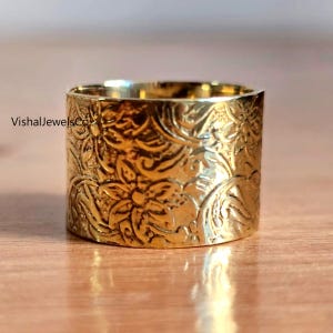 May include: A wide, gold-colored ring with an embossed floral pattern. The ring has a cylindrical shape and a polished finish. The ring is likely made of metal and is displayed on a wooden surface.