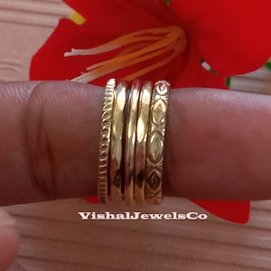 Puede incluir: Un conjunto de anillos dorados, cada uno con un diseño único, incluyendo bandas texturizadas y estampadas. Los anillos se apilan en un dedo, mostrando sus diversos estilos. La imagen incluye el texto "VishalJewelsCo".