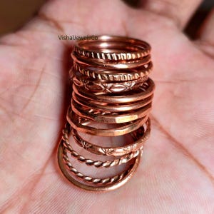 May include: A collection of copper-coloured rings with diverse textures, including smooth, patterned, and twisted designs. The rings are stacked, displaying the different styles. The rings are a warm, metallic colour.