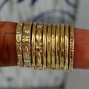 Op de afbeelding: Een verzameling goudkleurige ringen, sommige met geëtste bloemen- en geometrische patronen, gestapeld op een vinger. De ringen variëren in ontwerp, met sommige gladde banden en andere met een textuuroppervlak. De ringen worden in een close-up getoond.