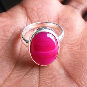 May include: A silver ring featuring a large, polished, oval-shaped hot pink gemstone with subtle horizontal banding. The stone is set in a smooth, plain silver bezel, and the band is made of a polished silver metal with a simple, rounded design.