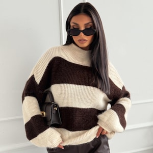 Puede incluir: Un suéter a rayas crema y marrón con cuello alto. El suéter tiene rayas horizontales anchas y mangas largas. Se ve un pequeño bolso negro.