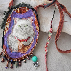 Puede incluir: Un collar con colgante hecho a mano con un retrato de gato. El colgante tiene un borde morado con una ilustración de gato, un collar rojo y ojos verdes. El collar incluye cuentas, una cuenta turquesa y un dije de flor plateada, con una correa de tela marrón y roja.