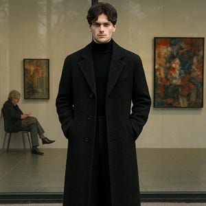 以下が含まれることがあります： 黒いオーバーコートと黒いタートルネックを着た人がガラスの壁の前に立っています。長いコートは質感があり、ボタンで留められています。その人は黒いズボンと靴を履いています。2つの絵が壁に掛かっています。