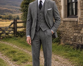 May include: A three-piece grey tweed suit comprising a single-breasted jacket, a matching button-up waistcoat, and tailored trousers. The outfit is worn with a crisp white collared shirt, a solid black necktie, and a white pocket square in the jacket.