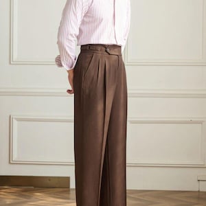 May include: A light pink and white striped dress shirt paired with high-waisted, pleated brown trousers. The outfit is completed with black leather loafers. The shirt has a classic collar and buttoned cuffs.