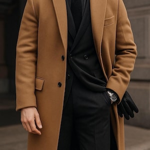 May include: A tan overcoat with a black suit, shirt, and tie. The coat has a classic design with a notched lapel and two front pockets. The person is wearing black gloves and a watch.