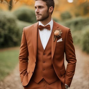 Könnte beinhalten: Ein dreiteiliger Anzug in Rostbraun, bestehend aus Jacke, Weste und Hose, kombiniert mit einer passenden Fliege und einem weißen Hemd. Eine florale Knopflochblume und ein Einstecktuch setzen Akzente. Der Mann steht im Freien.