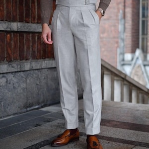 May include: Light gray pleated trousers with a high waist and a belt. The trousers are paired with brown leather tassel loafers. The person is wearing a brown long-sleeve shirt. The trousers have a cuff at the bottom.
