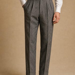 May include: High-waisted grey trousers featuring a textured herringbone pattern, double pleats at the front, and side buckle adjusters on the waistband. The trousers have a straight-leg cut with cuffed hems and are paired with a white button-down shirt.