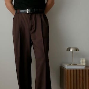 May include: A black short-sleeved t-shirt tucked into high-waisted, wide-leg dark brown trousers. The trousers feature a central pressed crease down each leg and are secured with a black leather belt featuring a silver-toned rectangular buckle.