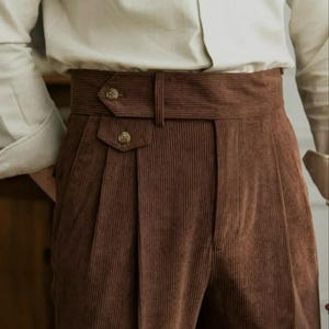 Pode incluir: Calças de veludo cotelê castanhas de cintura subida, com um cós largo, fecho de aba lateral, dois pequenos botões decorativos e pregas profundas na frente. As calças são combinadas com uma camisa de manga comprida de cor clara.