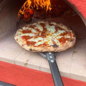 May include: A round pepperoni pizza with melted white cheese and red tomato sauce sits on a metal pizza peel inside a wood-fired oven. The oven has a bright red exterior and a stone base, with visible orange flames burning in the background.