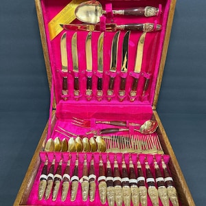 May include: A vintage silverware set in a wooden case. The set includes gold-colored knives, forks, and spoons with dark brown handles and decorative gold accents. The interior of the case is lined with pink velvet.