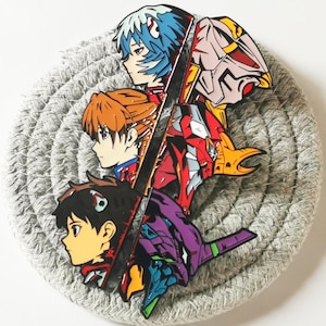 Może przedstawiać: Kolorowa przypinka przedstawiająca postacie z anime i robota, umieszczona na szarej, okrągłej podstawce. Przypinka przedstawia postacie z niebieskimi, pomarańczowymi i czarnymi włosami oraz czerwono-żółtego robota. Podstawka ma wzór plecionej liny.