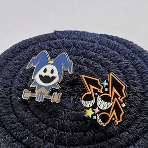 Könnte beinhalten: Zwei Emaille-Anstecknadeln auf einer dunkelblauen, strukturierten Oberfläche. Eine Nadel zeigt einen blau-weißen Narren-ähnlichen Charakter mit japanischem Text. Die andere ist ein schwarz-oranges Design mit einem lächelnden Gesicht und Sternakzenten.